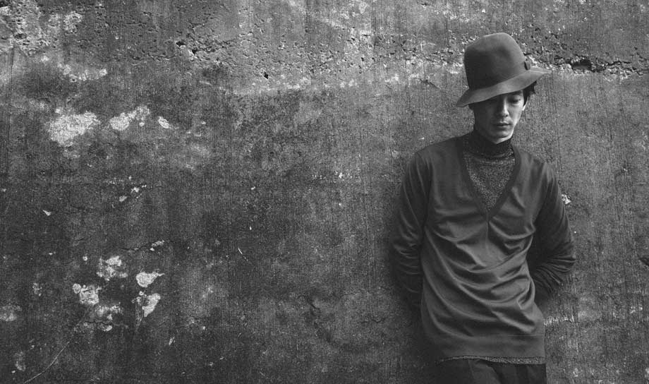 Ryo leaning against a concrete wall, wearing a hat by Margaret Howell, a lurex polo shirt by Prada and trousers by J.Lindeberg